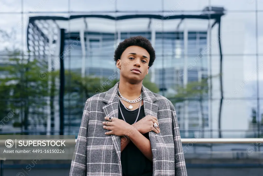 Confident young African American male teenager model in trendy tartan coat and stylish accessories looking at camera while standing against contemporary building with glass wall