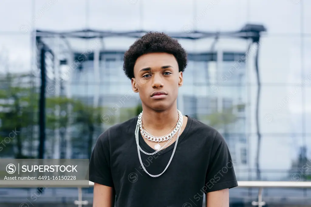 From below of young curly haired African American teenage guy in black t shirt and trendy neck chains looking at camera while standing against blurred modern building with glass wall