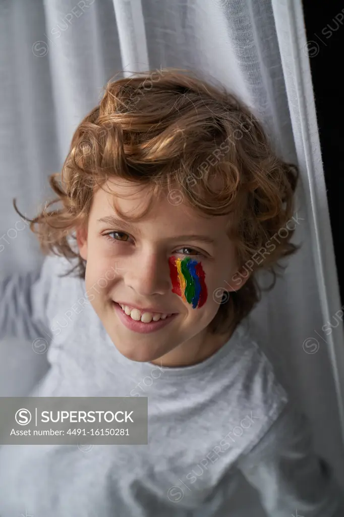 From above cheerful boy with rainbow under eye smiling looking at camera at home during quarantine