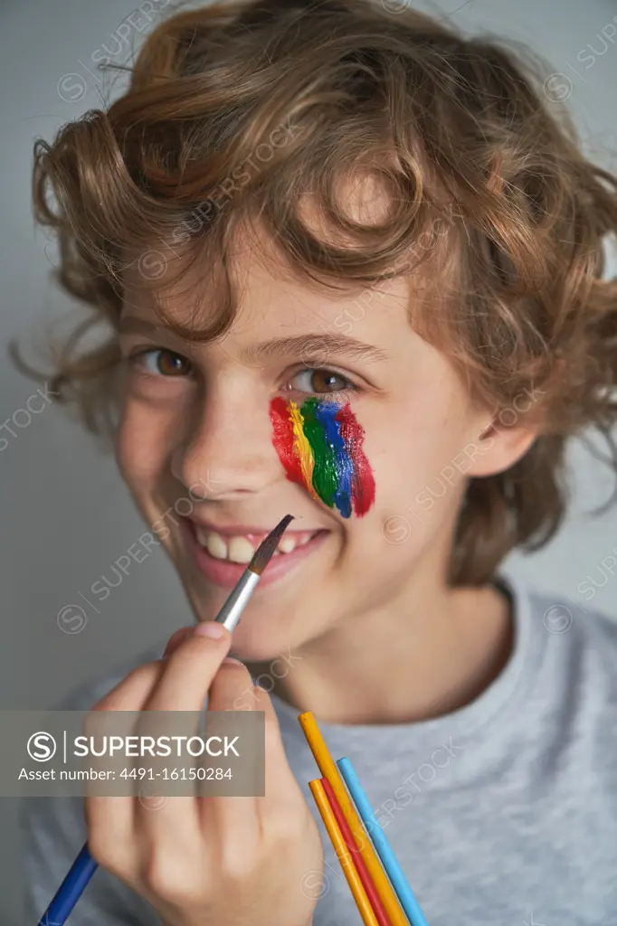 Happy boy painting colorful rainbow under eye and looking at camera while staying home during pandemic