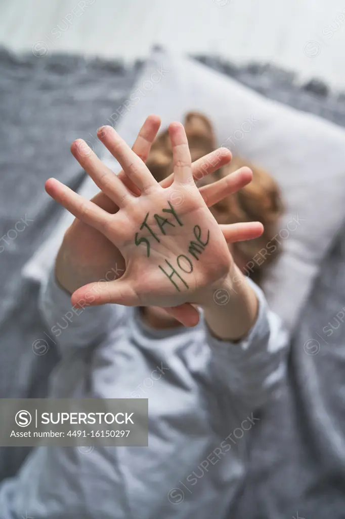 From above unrecognizable boy showing hand with Stay Home writing while lying on blanket and pillow during pandemic