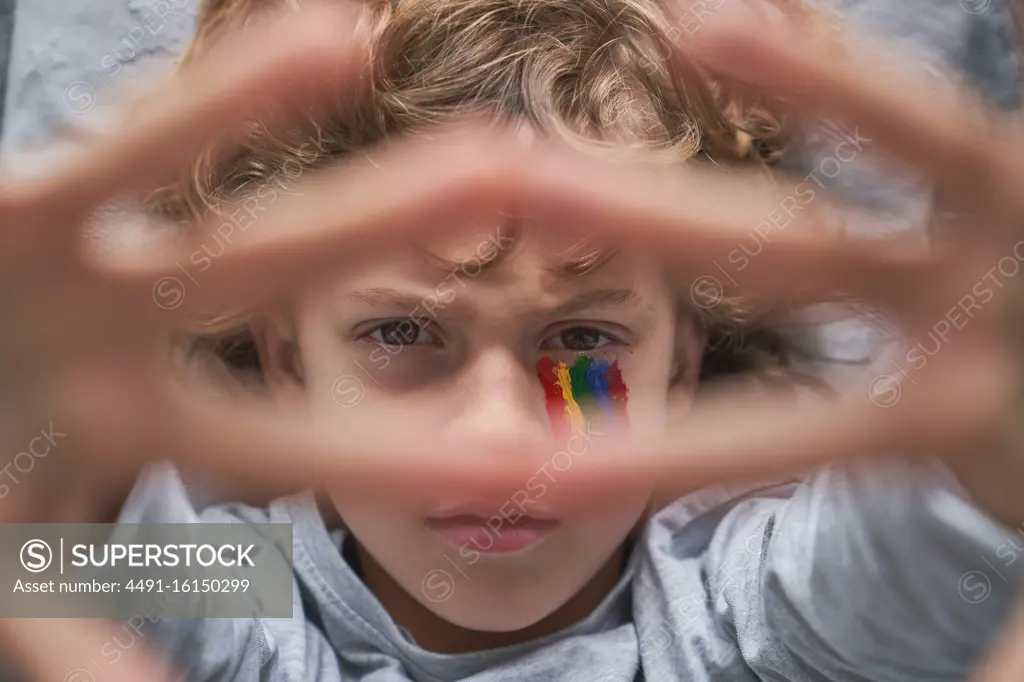 Top view of unhappy boy with colorful rainbow under eye showing stop gesture with hands with stay home inscription to the camera while lying on pillow and blanket on floor