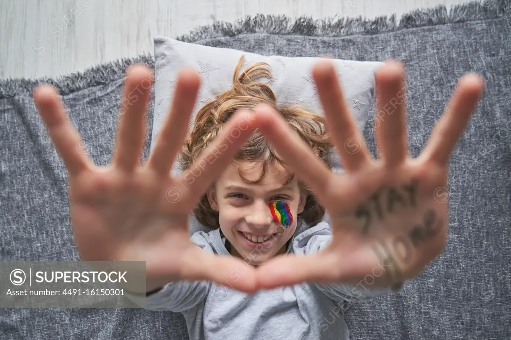 Top view of happy boy with colorful rainbow under eye showing stop gesture with hands with stay home inscription to the camera while lying on pillow and blanket on floor