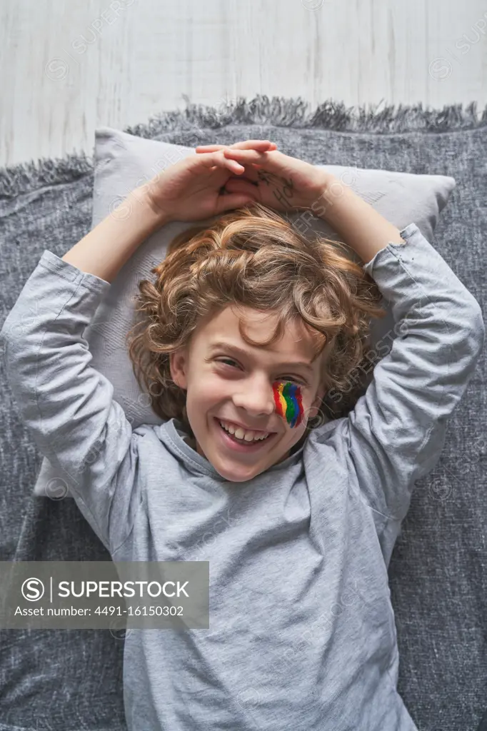 Top view of happy boy with rainbow under eye lying on blanket and pillow and laughing while staying home during quarantine