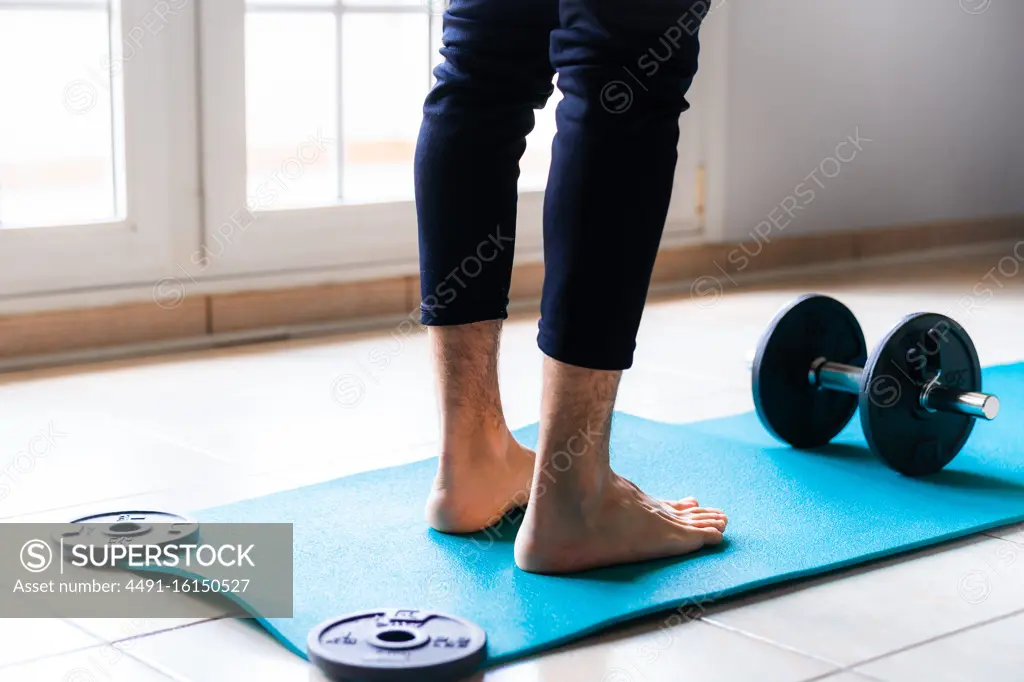 Low angle back view of crop barefooted male athlete in sportswear standing on bright blue sports mat against black collapsible dumbbell while preparing to workout in light spacious gym