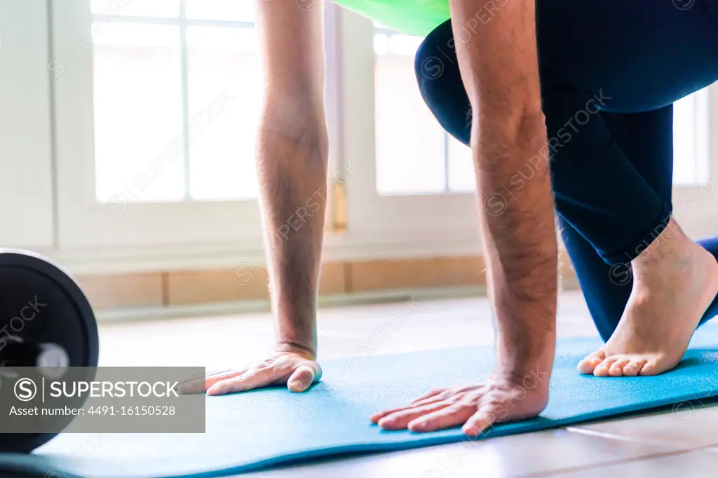 Low angle of unrecognizable determined male athlete in active wear warming up alone on vivid blue sports mat near black collapsible dumbbell at home