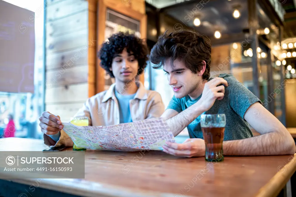 Multiethnic young homosexual men looking at direction navigation map and fresh drinks smiling while sitting at cafe table during romantic date