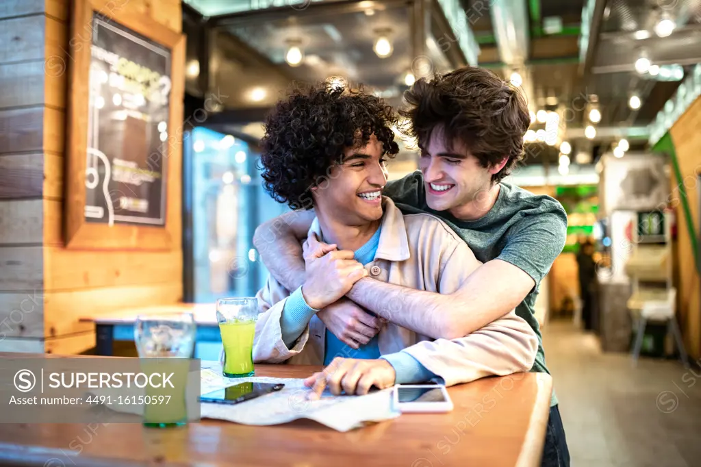 Multiethnic young homosexual men embracing each other looking at direction navigation map and having fresh drinks smiling while sitting at cafe table during romantic date