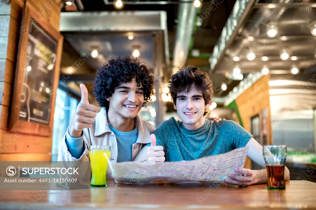 Multiethnic young homosexual men with direction navigation map and fresh drinks smiling and showing thumb up gesture while sitting at cafe table during romantic date