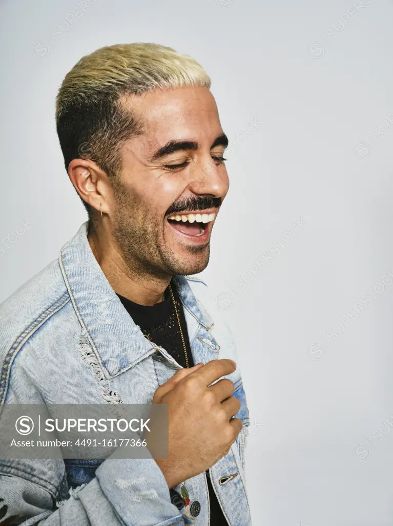 Side view of young unshaven stylish guy in trendy denim jacket laughing with closed eyes while standing against gray background