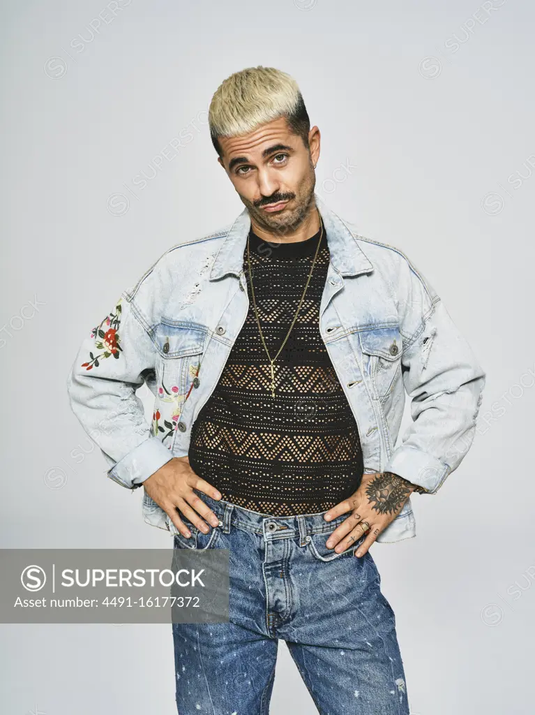 Young ethnic man making grimace face looking at camera wearing trendy denim jacket with floral pattern while standing against gray background