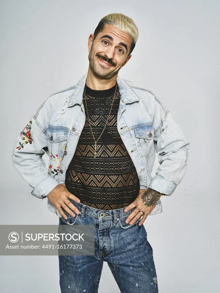 Young ethnic man making grimace face looking at camera wearing trendy denim jacket with floral pattern while standing against gray background
