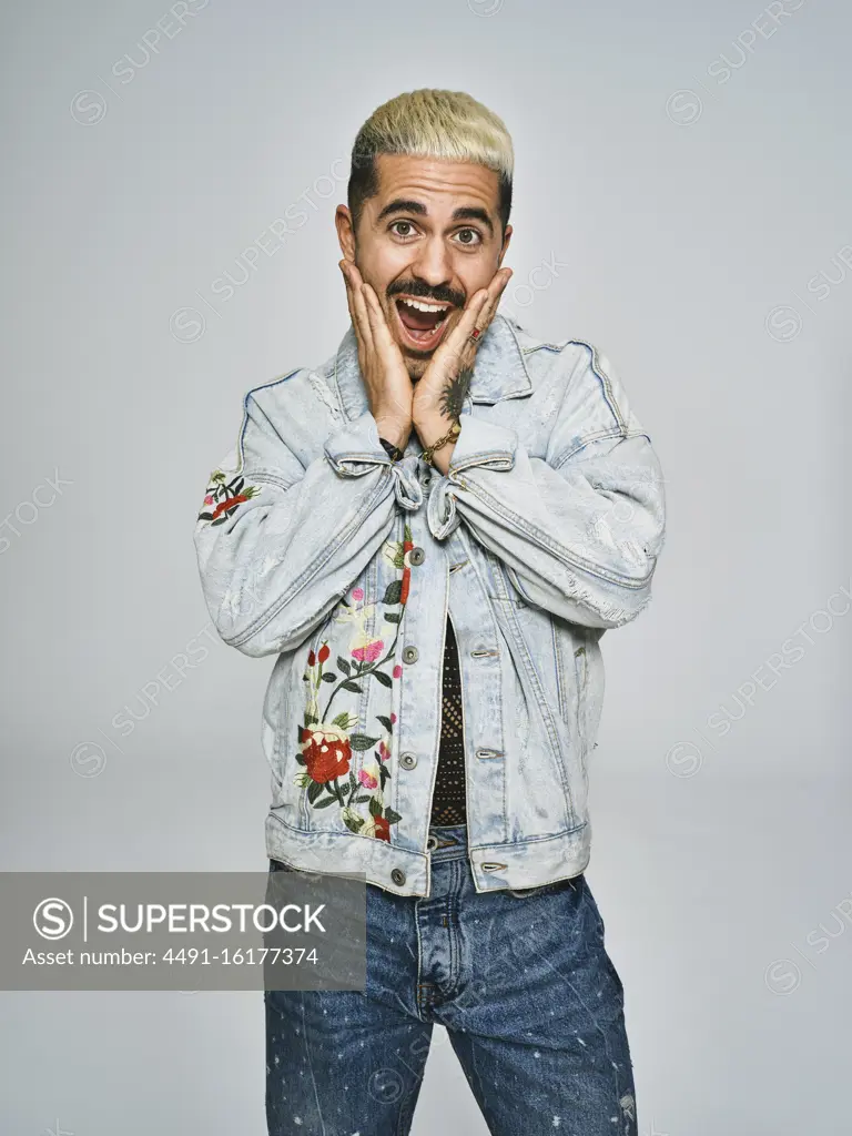 Young ethnic man making grimace surprised face looking at camera wearing trendy denim jacket with floral pattern while standing against gray background