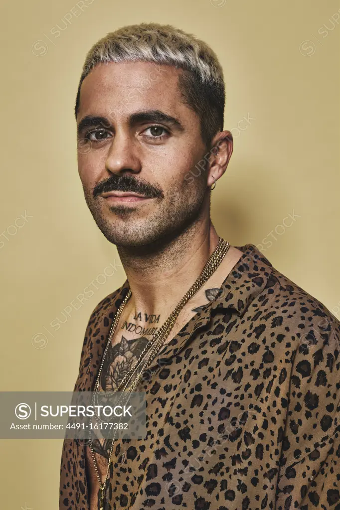 Portrait of cheerful fashionable male model with tattoos wearing trendy leopard shirt standing against beige background and looking at camera