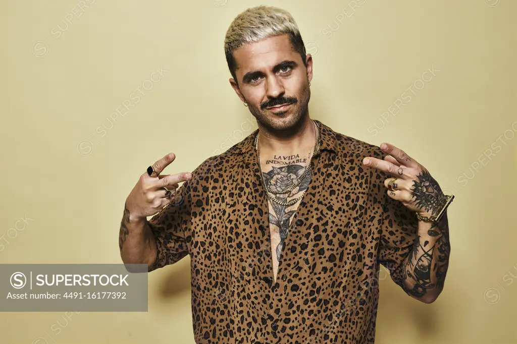 Positive young bearded guy in stylish leopard shirt revealing tattooed torso showing peace gesture while standing against beige background