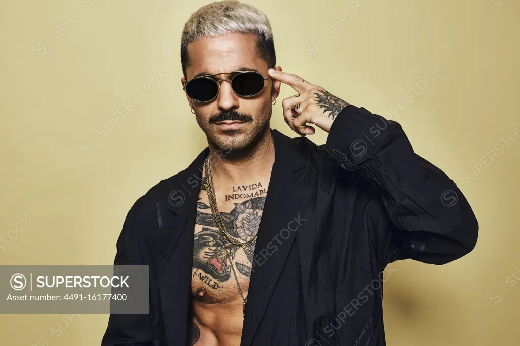 Portrait of brutal muscular sexy fit male with tattooed torso wearing black coat and stylish sunglasses and accessories standing against beige background looking at camera pointing finger to temple