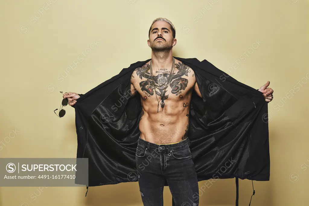 Brutal muscular sexy fit male opening with hands black coat showing up naked tattooed torso while on trendy jeans with stylish sunglasses standing against beige background looking at camera