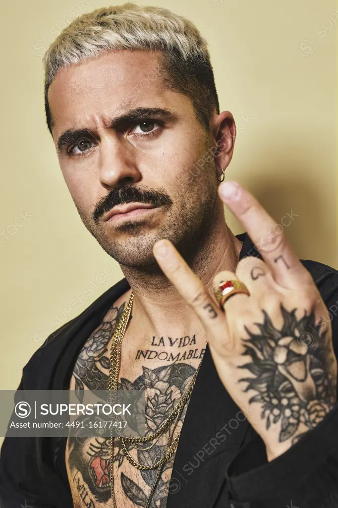 Young provocative male with mustache in black coat over naked tattooed torso showing rock sign gesture while standing against beige background looking at camera