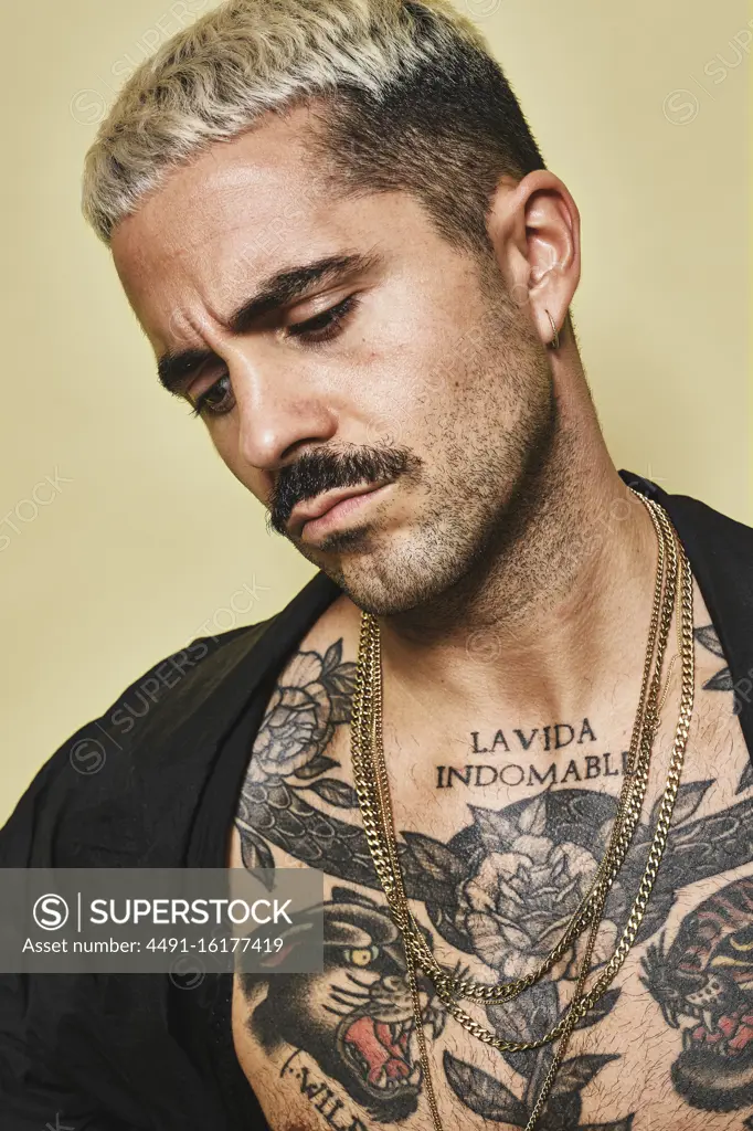 Confident arrogant stylish man with muscular tattooed torso wearing black coat looking away against beige background