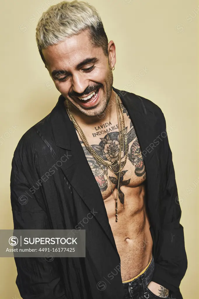 Confident cheerful stylish man with mustache showing off his muscular tattooed torso wearing black coat looking down against beige background