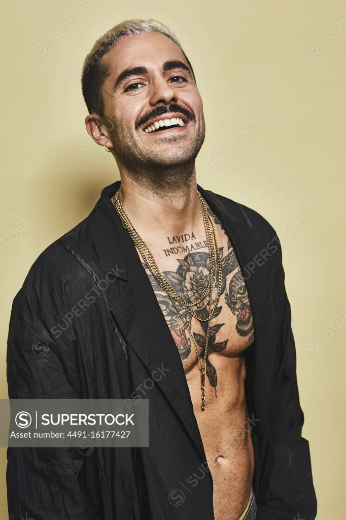 Confident cheerful stylish man with mustache showing off his muscular tattooed torso wearing black coat looking at camera against beige background