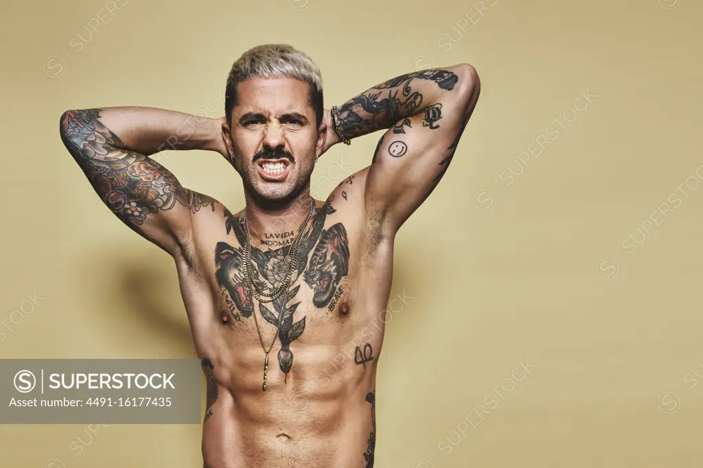 Handsome sexy attractive muscular male with various tattoos on naked torso and arms looking at camera making faces with mouth while standing against beige background