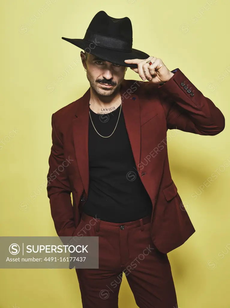 Handsome bearded well dressed male in trendy vinous suit and hat looking at camera against yellow background