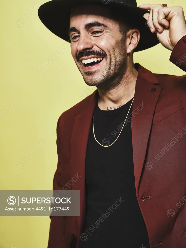 Laughing adult ethnic male in trendy vinous suit and hat looking at camera while standing against yellow background