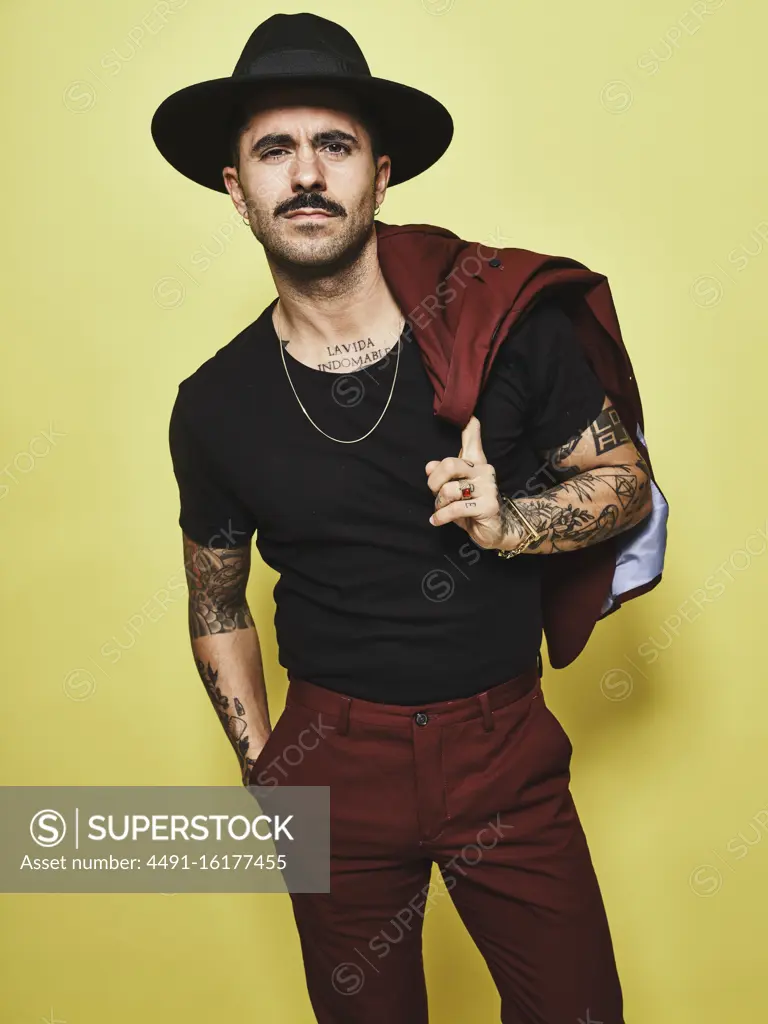 Handsome well dressed male with mustache in trendy hat and vinous suit holding suit coat over the shoulder looking at camera while standing against yellow background