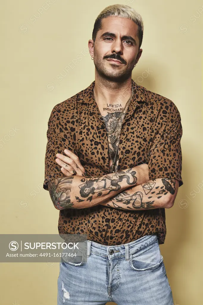 Fashionable male model with tattoos wearing trendy leopard shirt and jeans standing against beige background and looking at camera with arms crossed