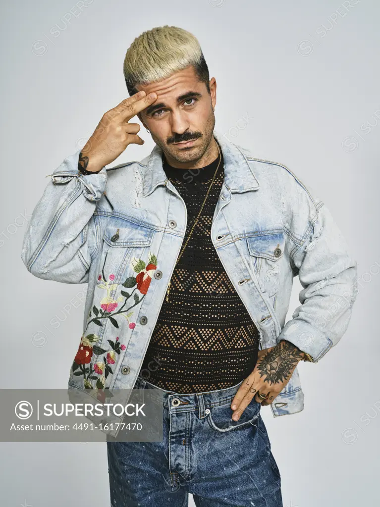 Young ethnic man making grimace doubting face with finger looking at camera wearing trendy denim jacket with floral pattern while standing against gray background