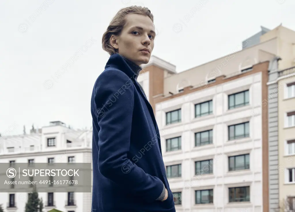 Side view of stylish young male with trendy hairstyle dressed in elegant coat standing against gray stone wall in city