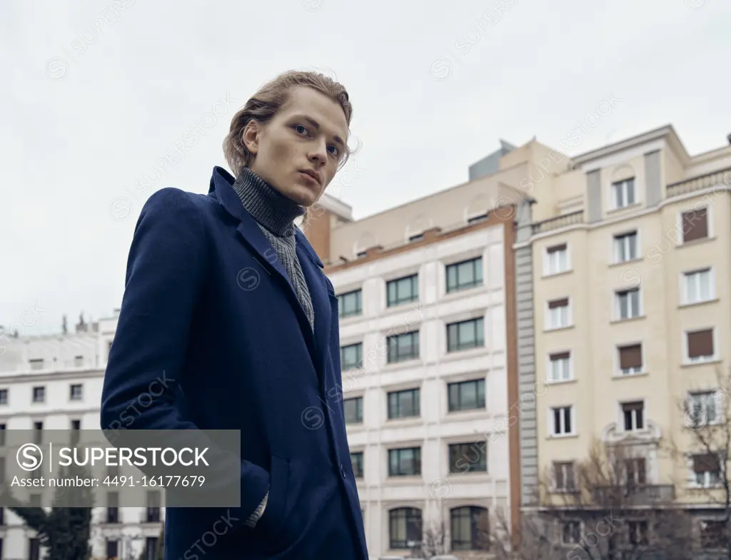 Side view of stylish young male with trendy hairstyle dressed in elegant coat standing against gray stone wall in city