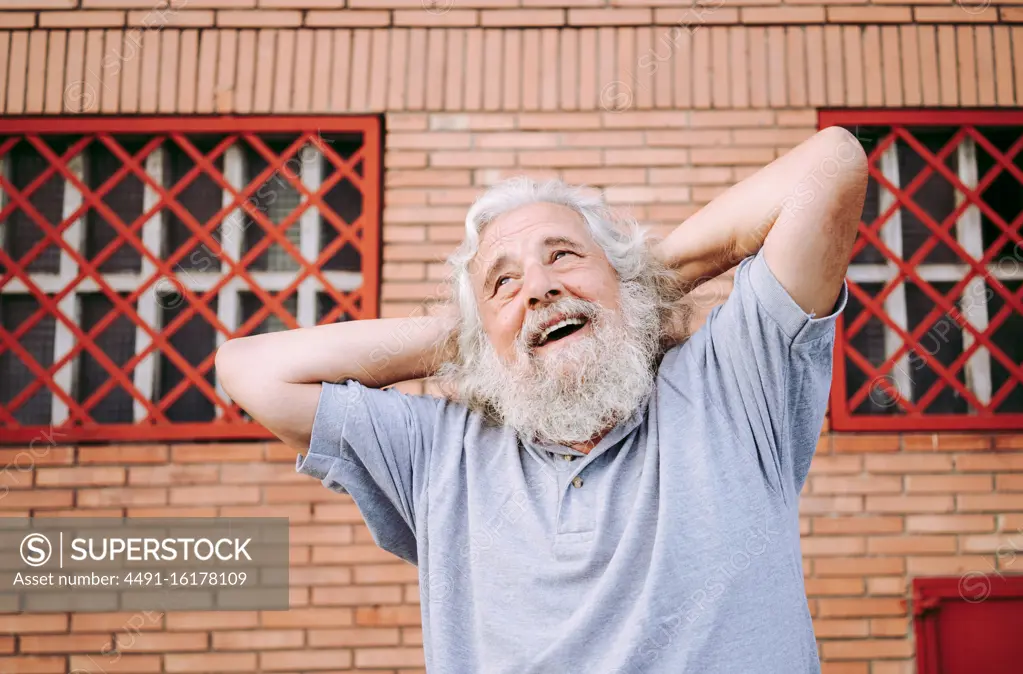 Aged bearded male in cotton tee shirt smiling and looking away while standing with hands behind head near loft building