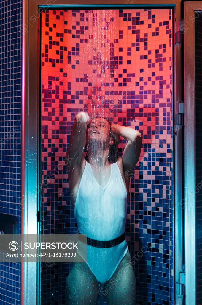 young female in swimwear enjoying water and relaxing in shower cabin illuminated with red and blue light