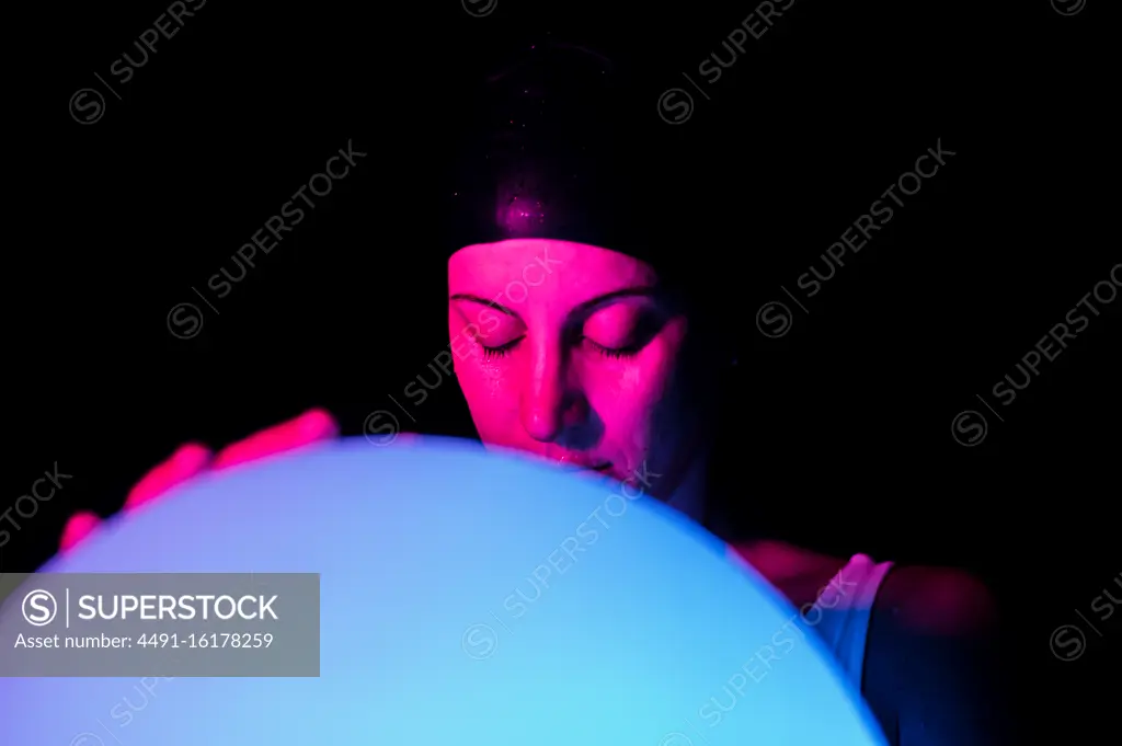 Tranquil young female in swimsuit and cap holding luminous ball while standing in dark swimming pool with neon light