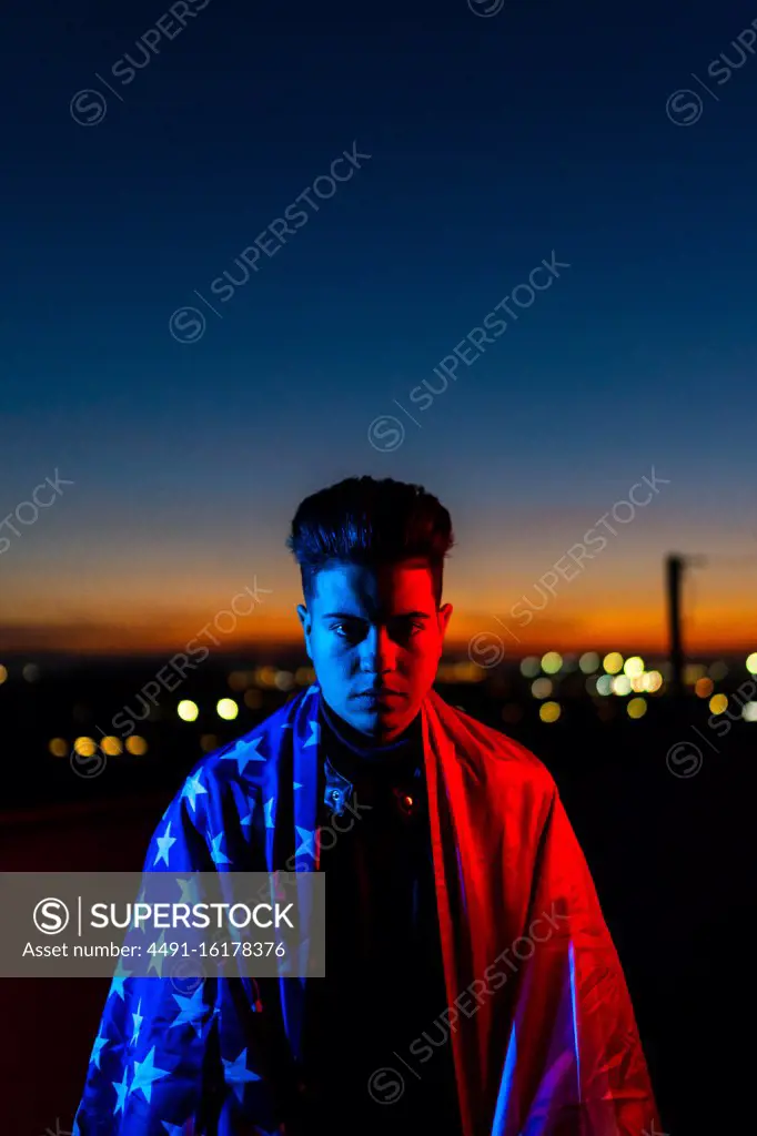 Serious young male in black leather jacket with American flag resting on shoulders looking at camera while standing in red and blue lights on dark street against sunset evening sky