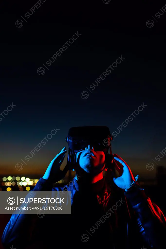 Modern male with VR headset exploring virtual world while standing in red neon light on street against dark sunset sky