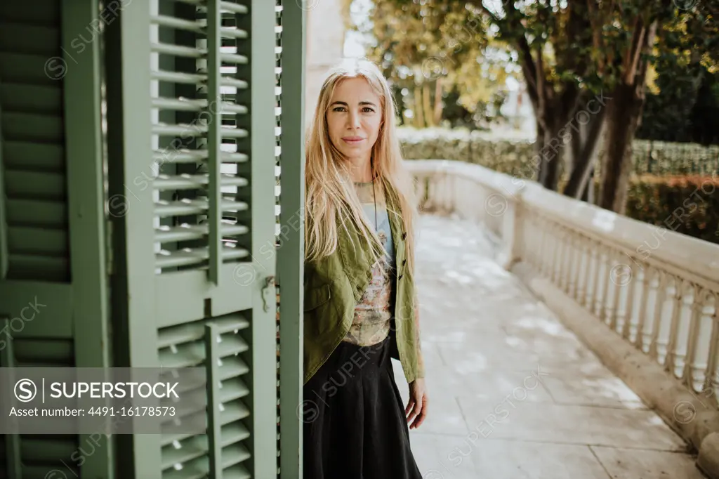 Adult female with blond hair looking at camera while standing near old building entrance on marble terrace on sunny day in garden