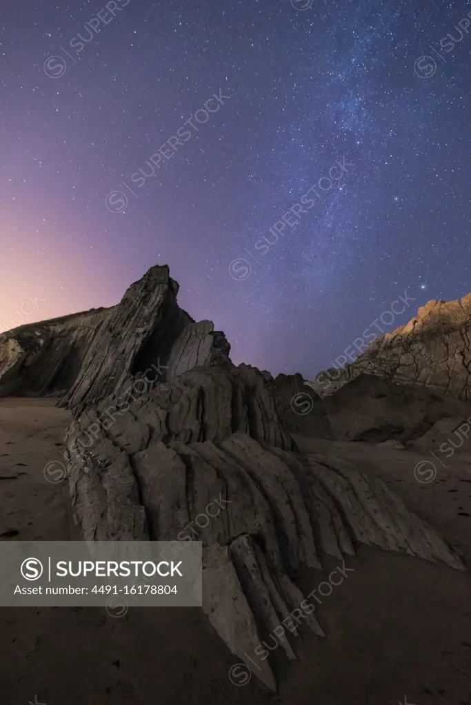 From below of rough cliff peak with colorful blue night sky and shiny stars on background