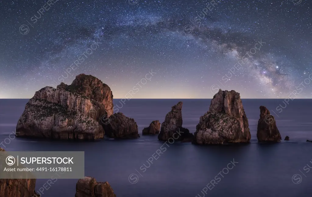 Big rough cliffs on blue calm ocean during bright evening under colorful starry sky with milky way