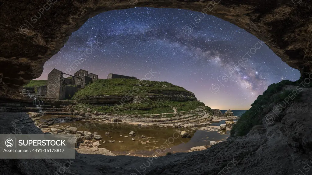 Stone rough cave on seashore with stone aged ruins and green hills under night sky with milky way and stars