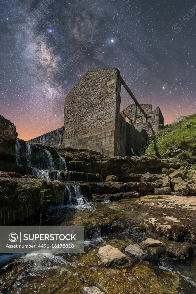 From below of ancient stone castle and small waterfall on stairs under dark sky with stars and milky way