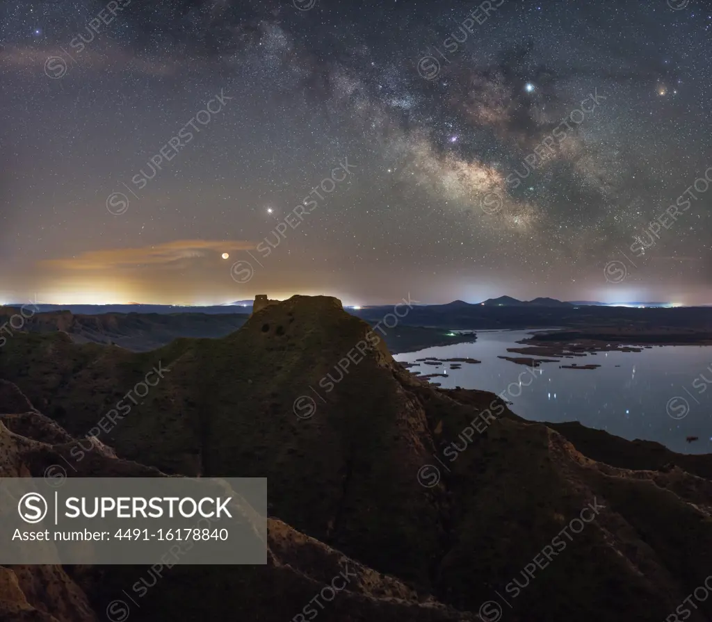 From above of tops of mountains and rocky valley with calm lake on background under colorful sky with milky way