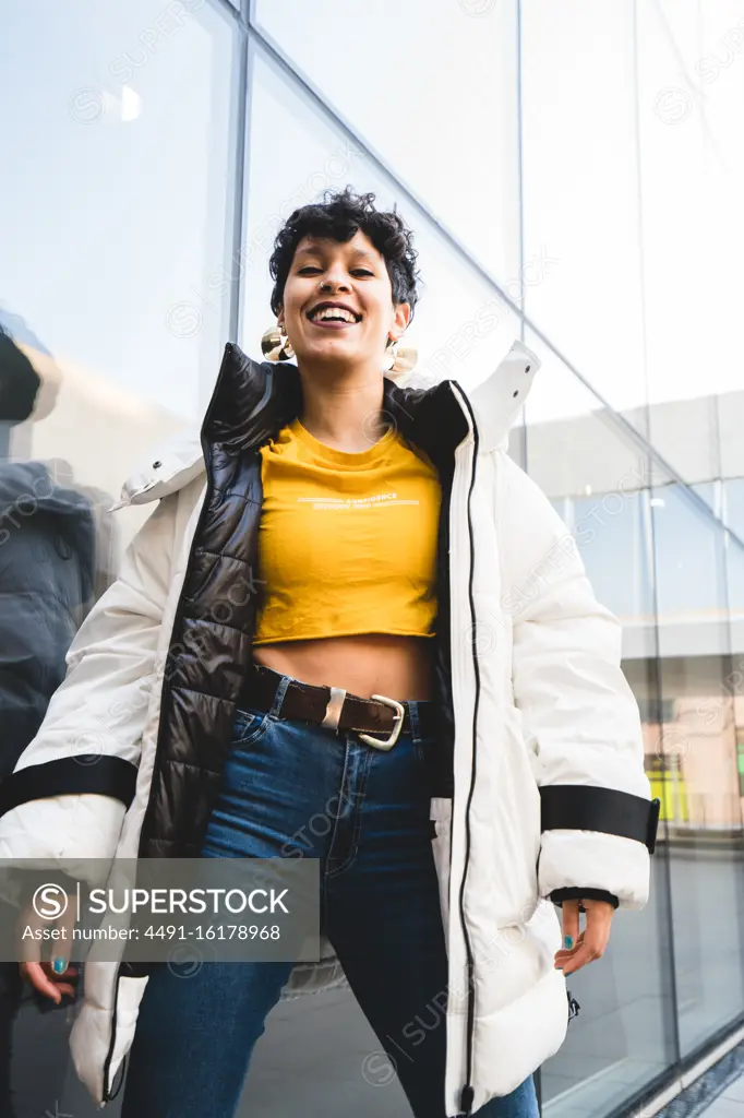 From below of joyful ethnic female teenager in colorful casual clothing and warm jacket laughing and looking at camera with modern building on background