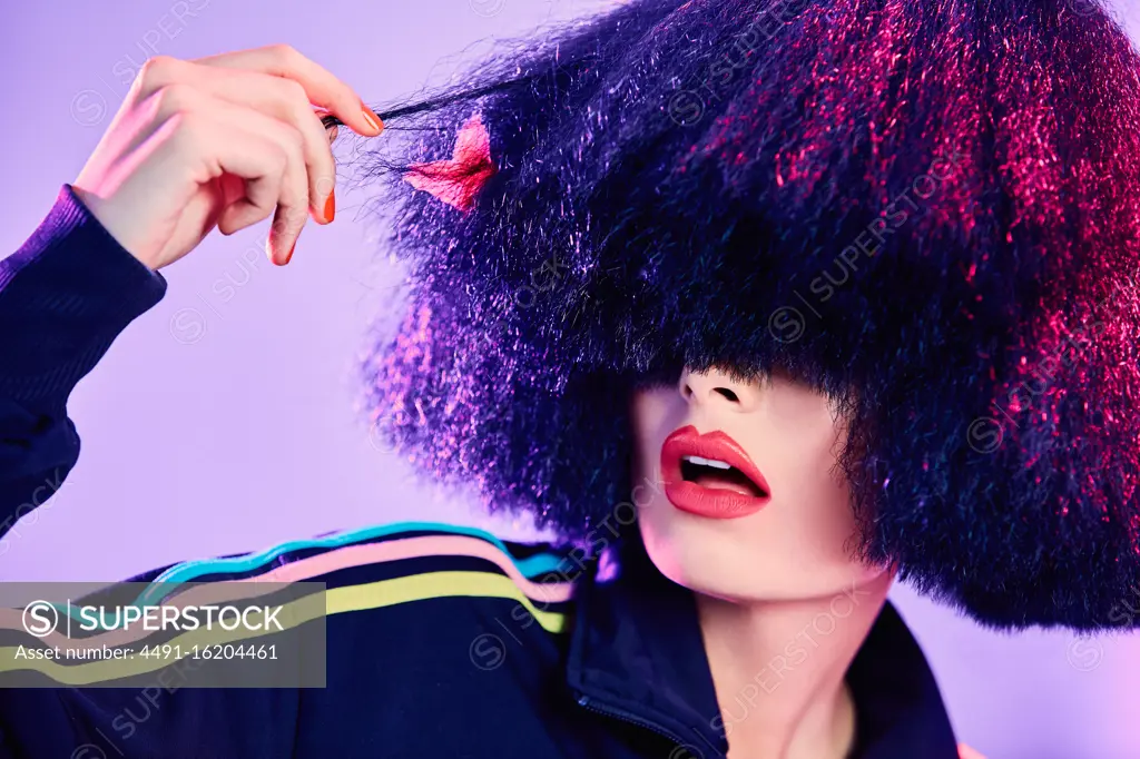 Astonished female in stylish sportswear inspecting fake hair under bright illumination against violet background