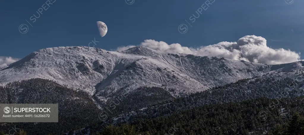 Breathtaking wide angle view of snowy mountain range against evening sky with white clouds and moon in nature
