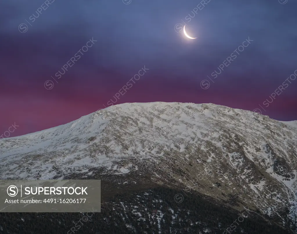 Breathtaking wide angle view of snowy mountain range against evening sky with white clouds and moon in nature