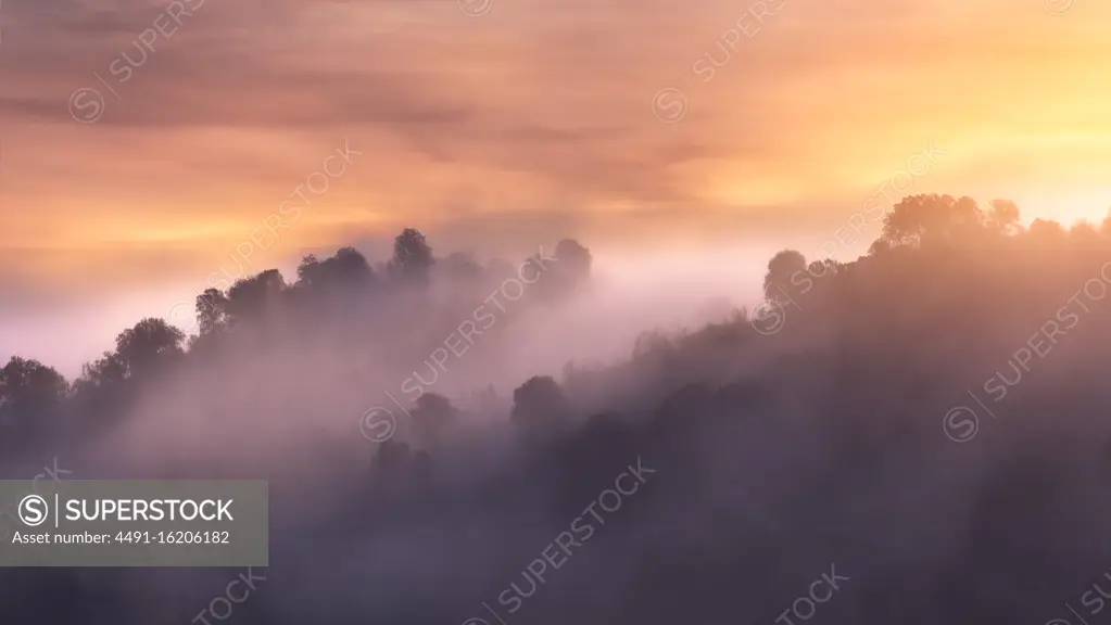 Rough mountain range with trees located against bright sunrise sky in hazy morning in nature