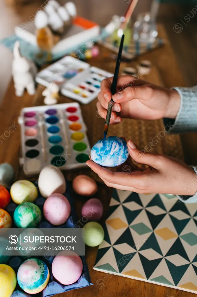Unrecognizable person in sweater covering fresh chicken eggs with blue paint while preparing for Easter celebration